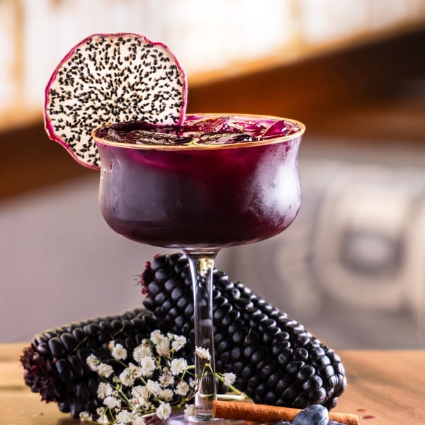 Purple cocktail with dragon fruit garnish, surrounded by black corn, spices, and blueberries.