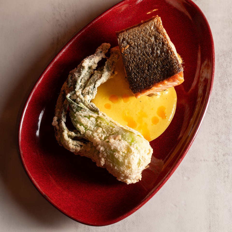 Crispy-skinned salmon with tempura-fried zucchini flower and yellow sauce on a red plate.