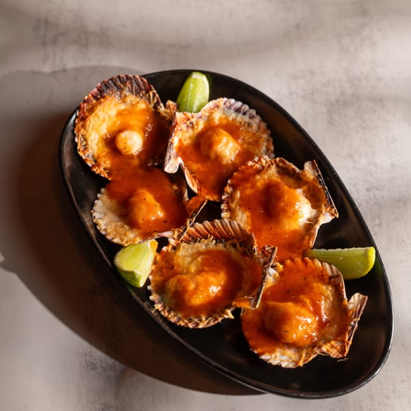 Six scallops in shells with orange sauce and lime wedges on a black plate.