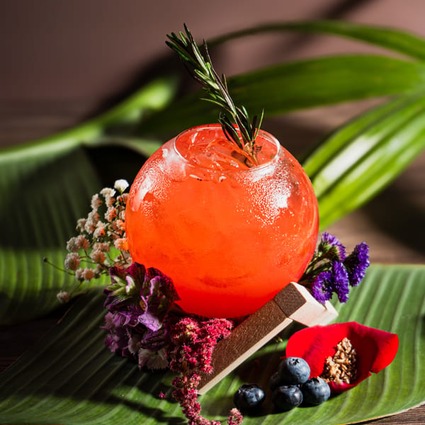 A vibrant red cocktail in a round glass, garnished with rosemary, flowers, and blueberries on a banana leaf.