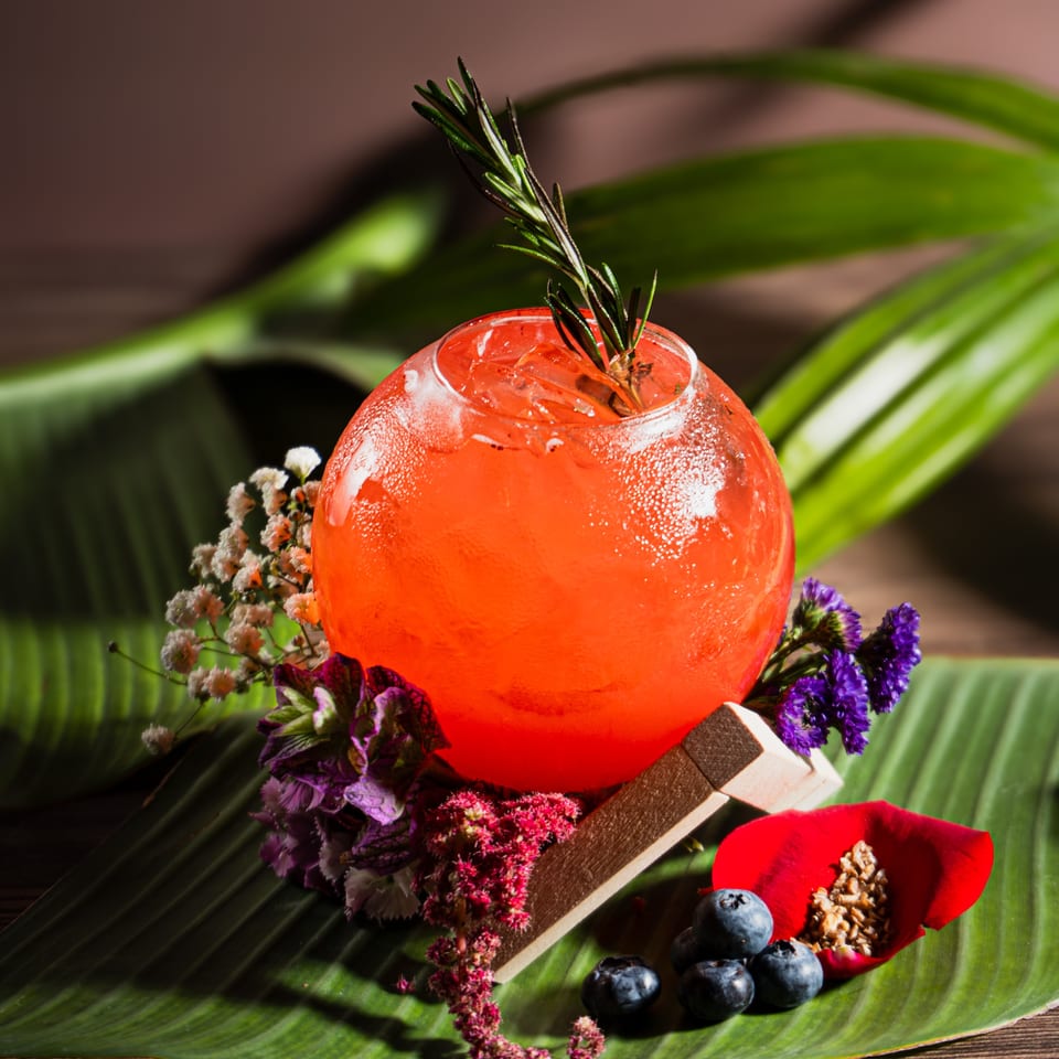 A vibrant red cocktail in a round glass, garnished with rosemary, flowers, and blueberries on a banana leaf.