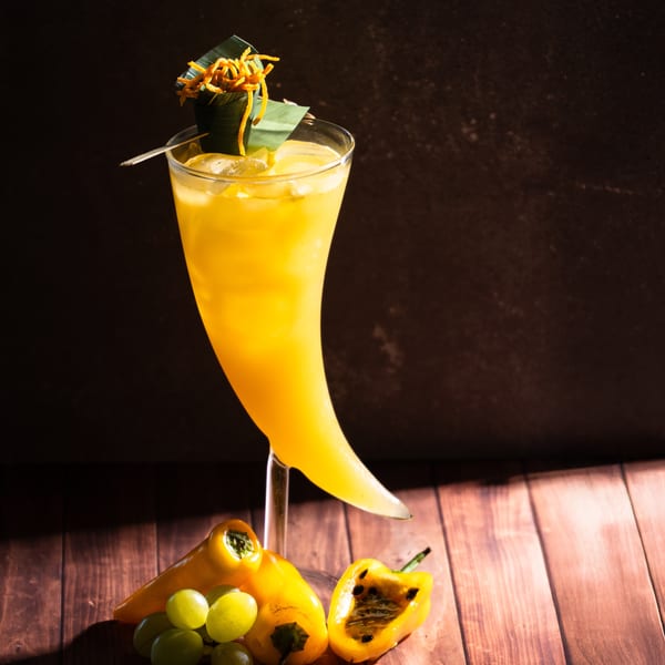 A vibrant yellow cocktail in a unique glass, garnished with orange peels and green leaves, alongside grapes and peppers.