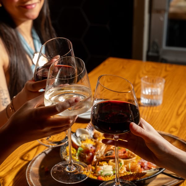 Three people clinking wine glasses over a paella dish with seafood on a wooden table.
