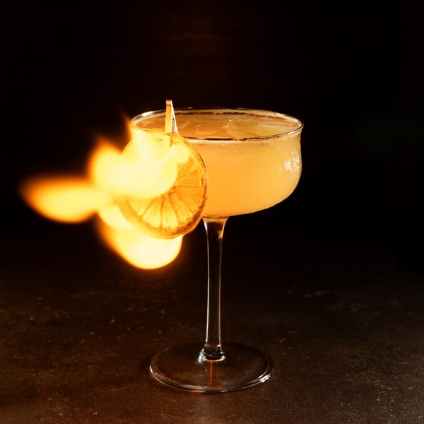 Flaming cocktail in a coupe glass, garnished with a dried orange slice, against a dark background.