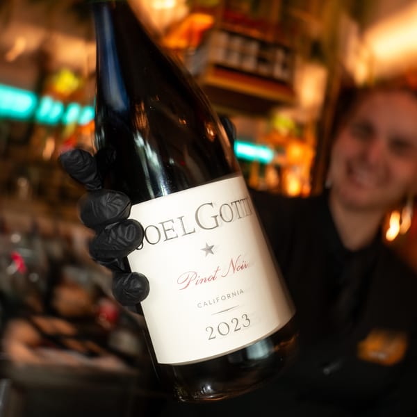 A gloved hand holds a bottle of Joel Gott Pinot Noir 2023, with a smiling server in the background.