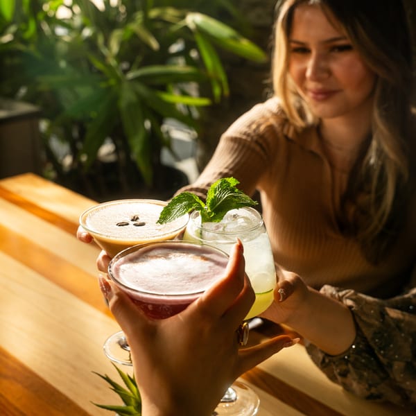 Two women clinking three cocktails, including an espresso martini and a mojito, at a sunny restaurant.