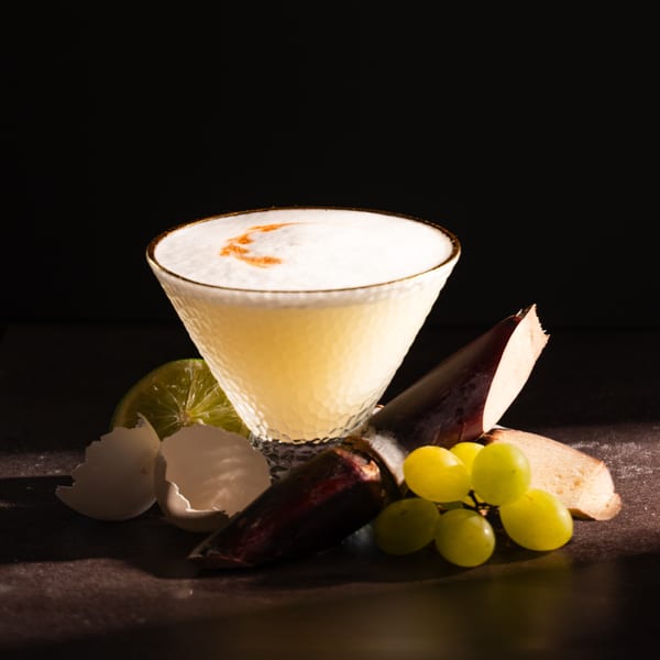 A pisco sour cocktail with eggshell, lime, sugarcane, and grapes on a dark surface.
