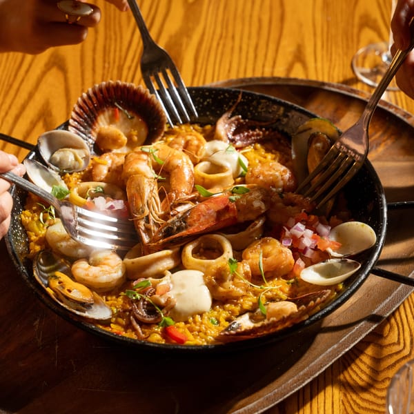 Hands with forks sharing a pan of seafood paella with shrimp, mussels, and squid on a wooden table.