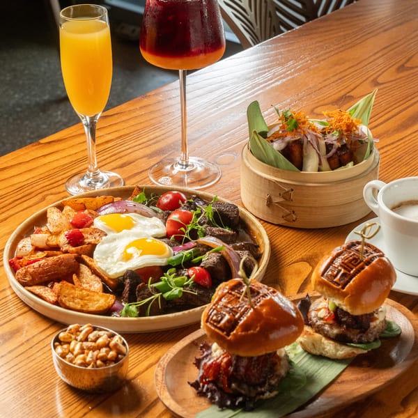 A brunch spread with steak and eggs, sliders, bao buns, and cocktails on a wooden table.