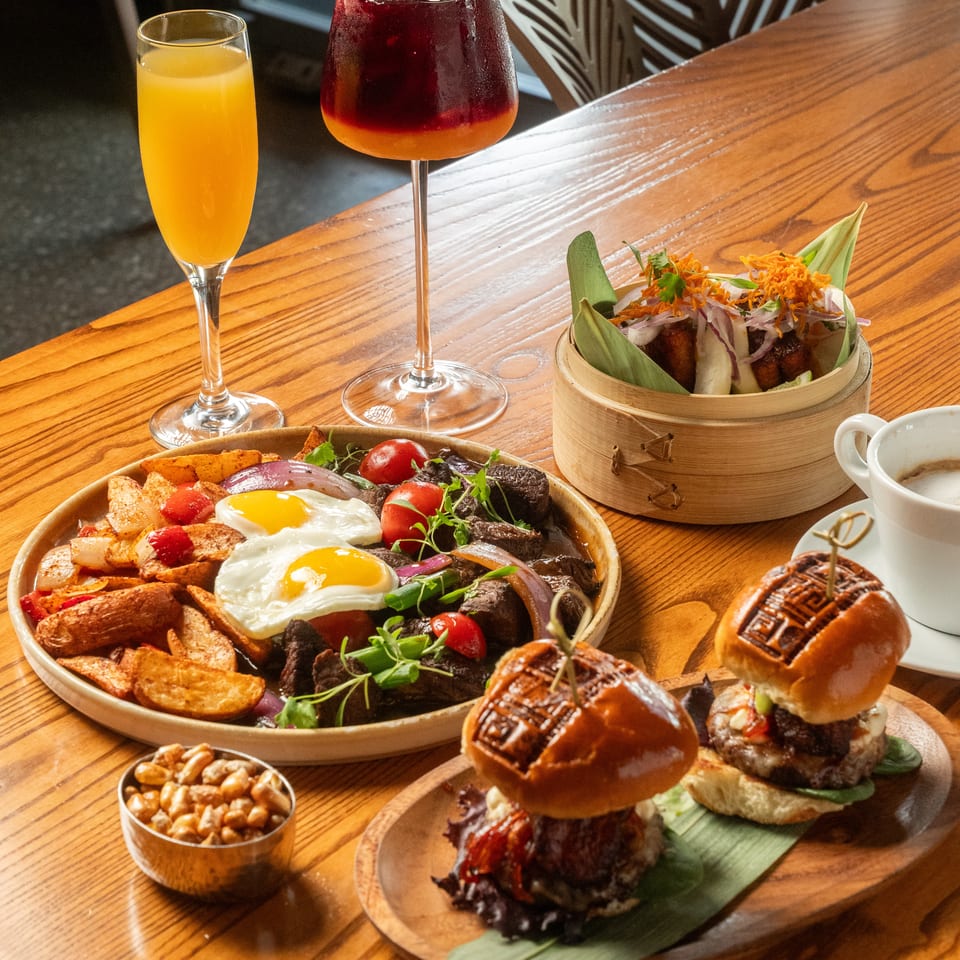 A brunch spread with steak and eggs, sliders, bao buns, and cocktails on a wooden table.