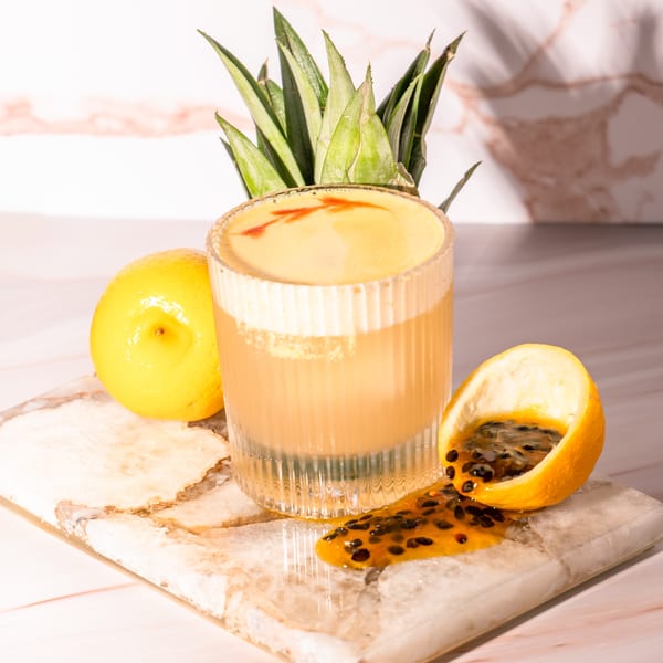 A cocktail with foam and red garnish, pineapple leaves, lemon, and spilled passion fruit on marble.