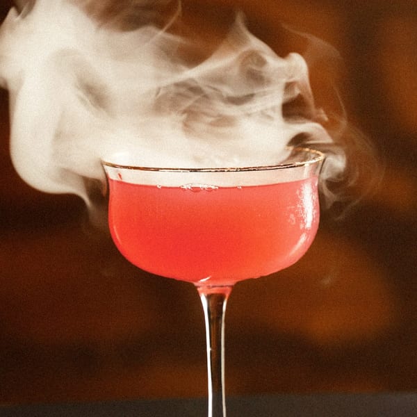 A vibrant pink cocktail in a coupe glass, emitting a plume of white smoke against a dark background.